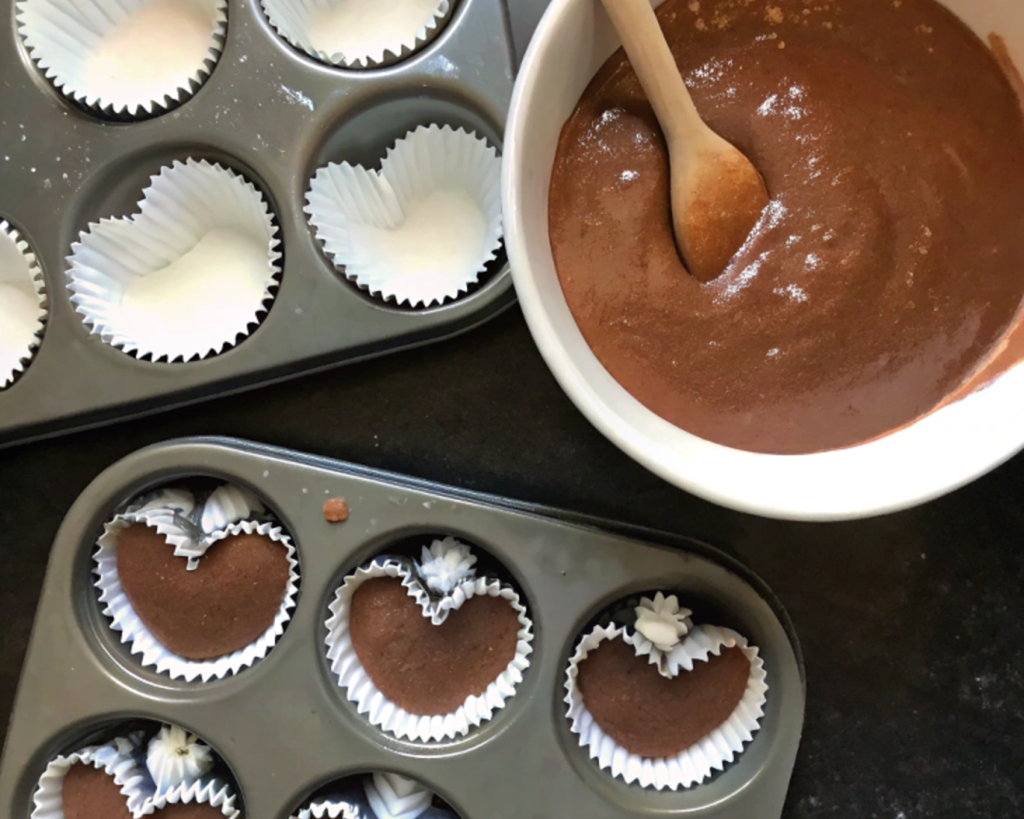 Heart-shaped chocolate cupcakes in a muffin tin, with chocolate batter in a white bowl and a wooden spoon beside it.