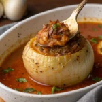 A close-up of a stuffed onion filled with a flavorful mixture, served in a rich tomato-based sauce with a spoon lifting the filling.