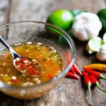 A bowl of homemade fish sauce recipe with chopped chili and garlic on a rustic wooden table, surrounded by fresh ingredients like lime and chili peppers.