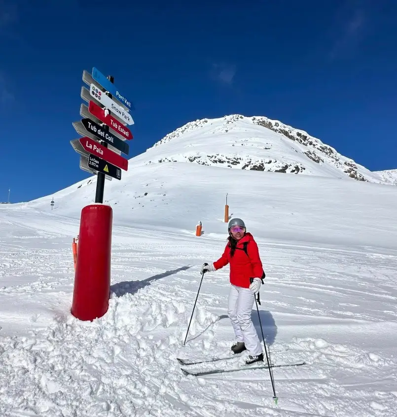 Saiko skiing in Andorra, wearing a red jacket and white pants, smiling under a bright blue sky with snowy mountains in the background
