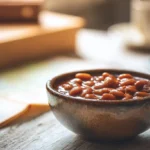 Rustic ceramic bowl filled with sweet and smoky baked beans without pork, set beside a cup of coffee and travel books in warm light.